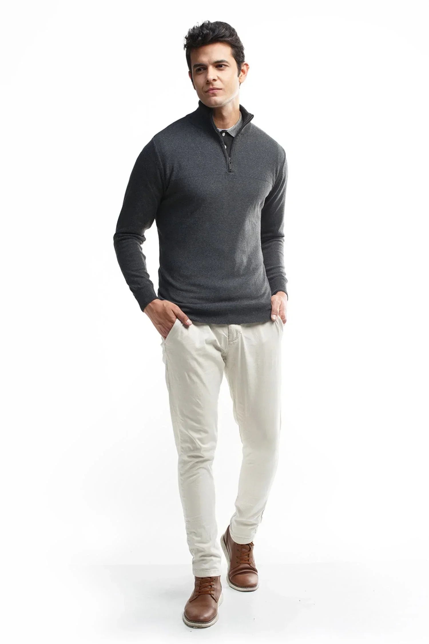 Hombre con suéter gris de punto, pantalón claro y botas marrones sobre fondo blanco.